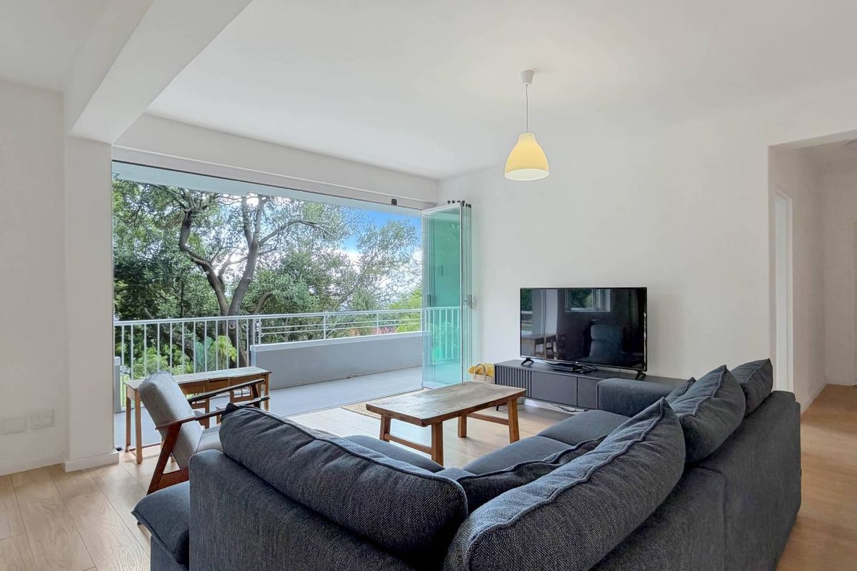 Bright living room featuring light wood-look flooring and large bi-fold doors opening onto a private balcony with leafy views.