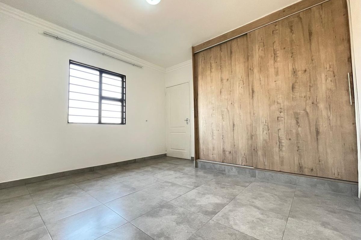 A clean, empty bedroom featuring modern grey floor tiles, a large built-in wooden sliding door wardrobe, and a window with security bars.