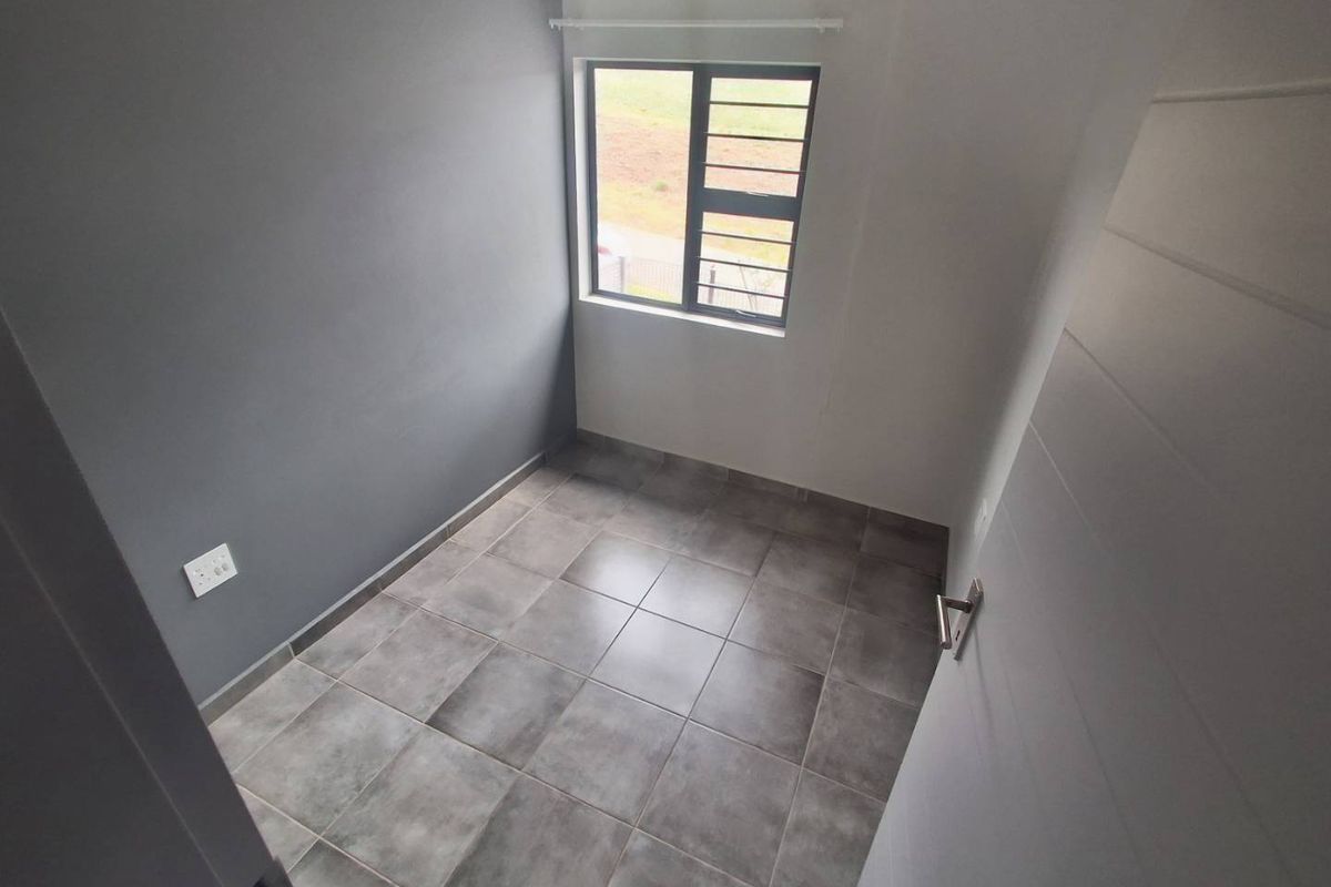 A clean, empty bedroom featuring modern grey floor tiles, a neutral color palette, and an aluminium window with security bars.