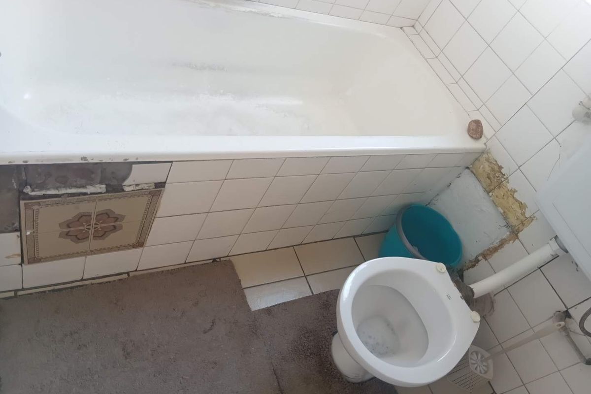 A dated bathroom featuring a standard bathtub and toilet, currently requiring significant repairs to wall tiling and surrounding finishes.