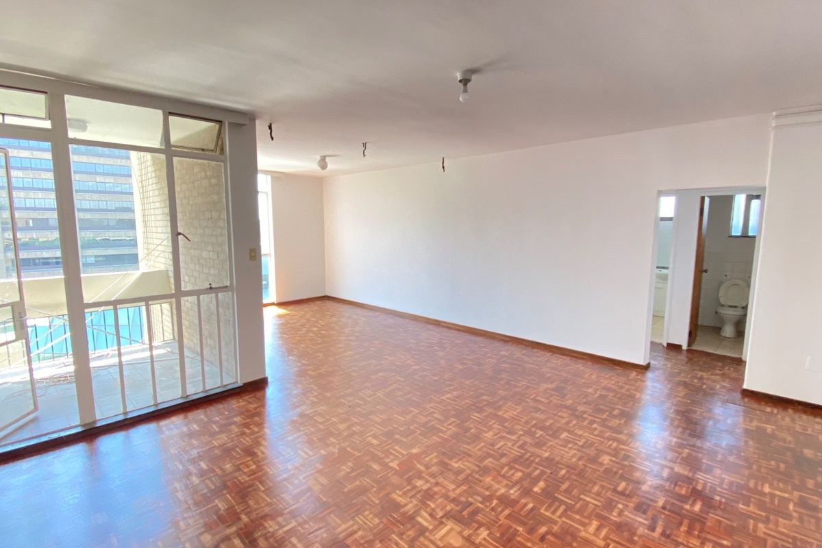Spacious, empty living area featuring classic parquet flooring, large glass sliding doors leading to a balcony, and direct access to an adjacent bathroom.