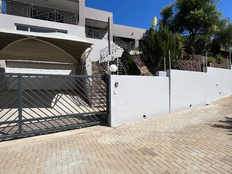 Front exterior view of a modern multi-level house featuring a paved driveway, secure electric gate, carport, and elevated balcony.