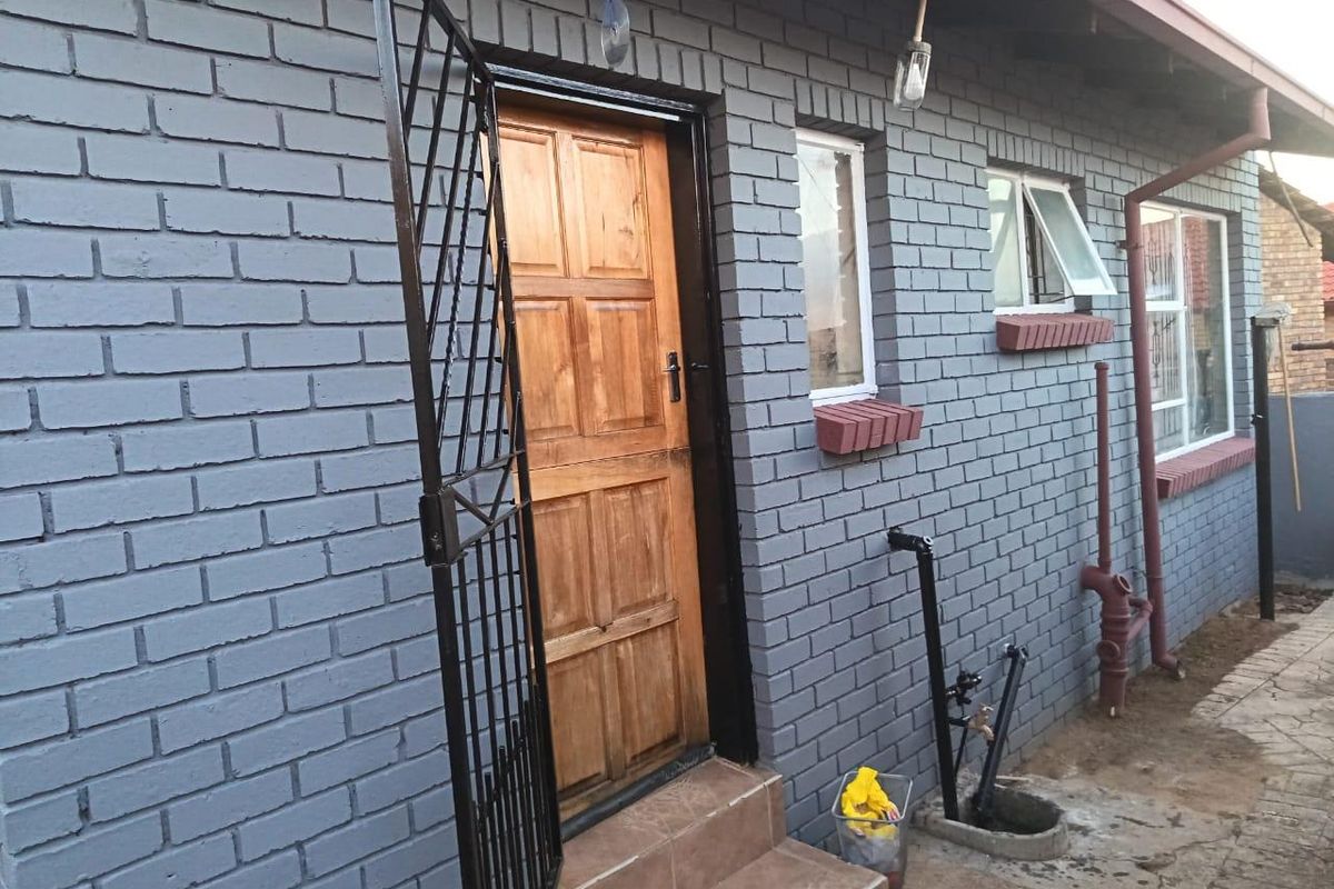Rear exterior view of the property featuring a painted brick facade, timber entrance door with security gate, and functional casement windows.