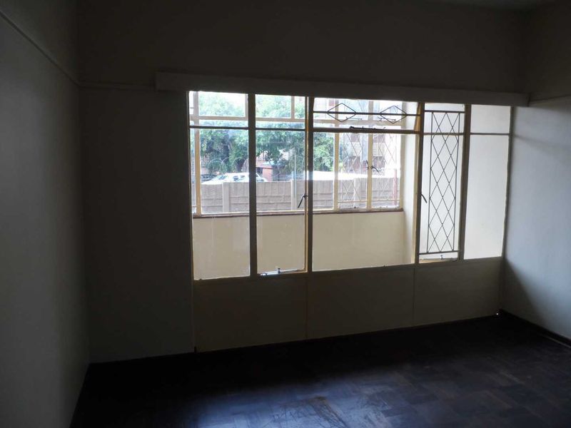 An empty bedroom featuring original parquet flooring and large steel-framed windows with security bars, offering a blank canvas for renovation.