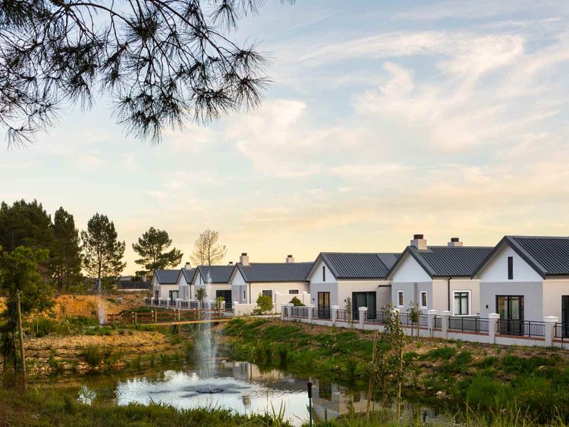 A scenic view of a row of modern residential units featuring metal roofs, overlooking a landscaped pond with a central fountain.