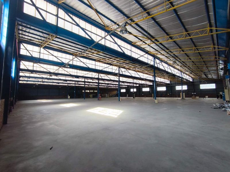 Expansive industrial warehouse space featuring high ceilings with exposed steel trusses, concrete flooring, and abundant natural light through translucent roof panels.
