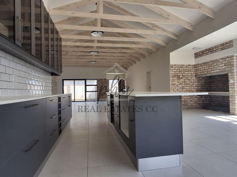 Spacious open-plan kitchen and living area featuring exposed timber trusses, a modern kitchen island, subway tile backsplash, and a feature brick fireplace.