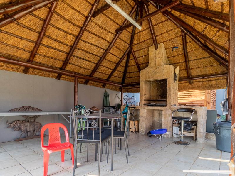 Covered outdoor patio area featuring a traditional thatched roof, exposed timber rafters, and a built-in stone braai fireplace.