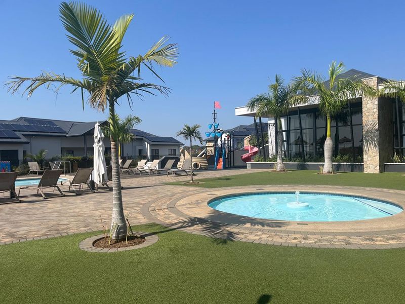 A well-maintained communal pool area featuring a fountain, paved patio, artificial lawn, and a children's playground set within a modern apartment complex.