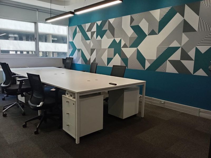 A modern office space featuring a large central desk, linear pendant lighting, carpeted flooring, and a decorative geometric accent wall.
