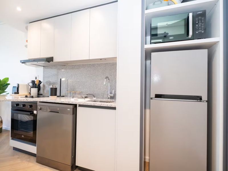 Modern compact kitchen featuring sleek white cabinetry, granite countertops, stone splashback, and integrated stainless steel appliances including an oven and dishwasher.