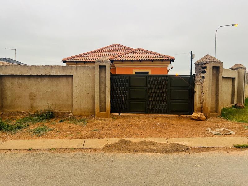 Front view of the property featuring a perimeter wall, metal sliding gate, and tiled roof structure with visible wear on the boundary pillars.