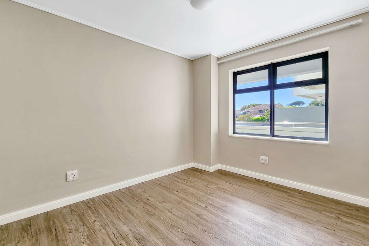 A bright, empty bedroom featuring modern laminate flooring, neutral wall tones, and a large aluminium-framed window providing natural light.