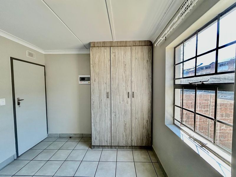 A clean, empty bedroom featuring neutral tiled flooring, built-in wooden cupboards, and a window fitted with security burglar bars.