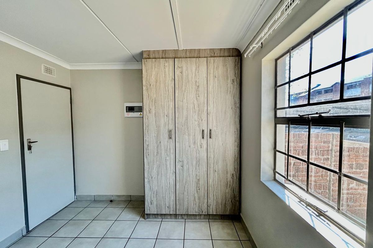 A clean, empty bedroom featuring neutral tiled flooring, built-in wooden cupboards, and a window fitted with security burglar bars.