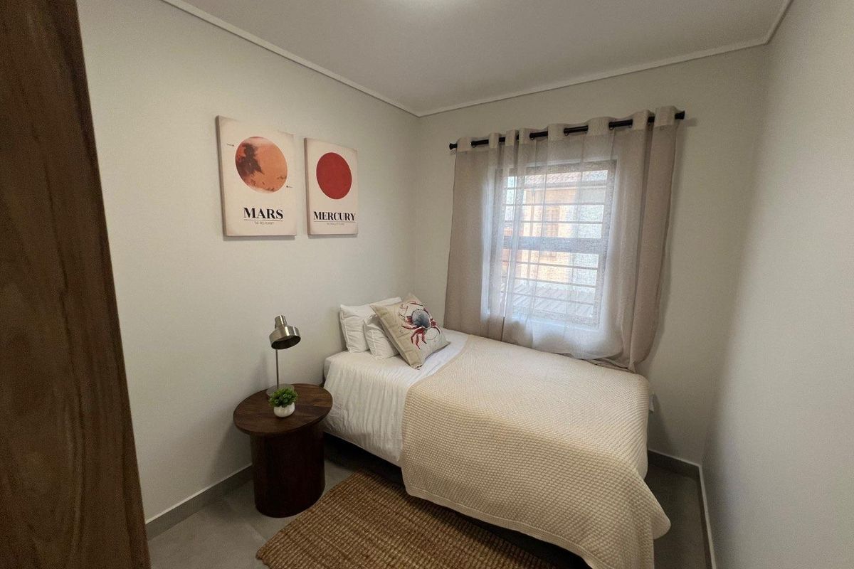 A compact bedroom featuring neutral wall tones, tiled flooring, and a window fitted with security bars and curtains.