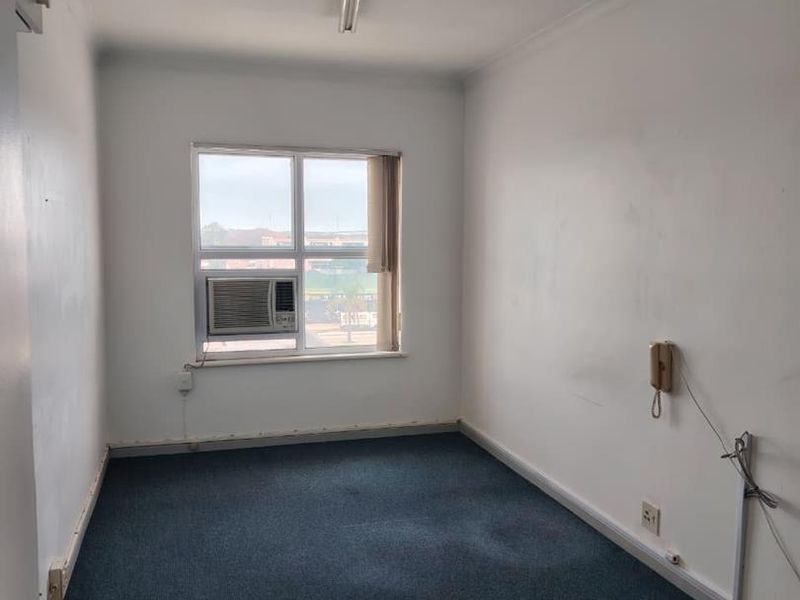 An empty office space featuring blue carpeting, a window-mounted air conditioning unit, and basic fluorescent lighting, suitable for a professional workspace.