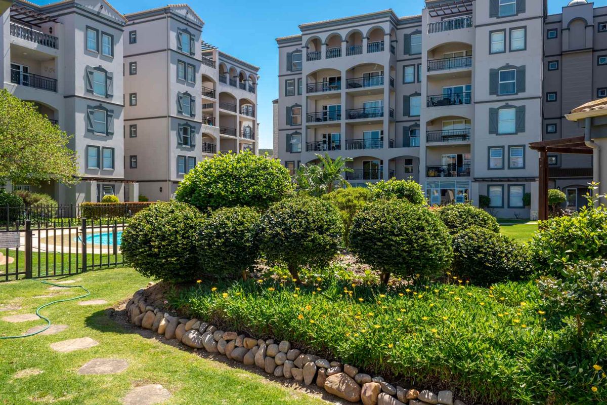 A well-maintained apartment complex exterior featuring landscaped gardens, stone retaining walls, and private balconies overlooking a communal outdoor area.