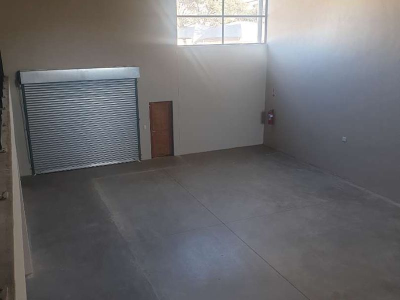 Spacious industrial warehouse unit featuring high ceilings with exposed steel trusses, concrete flooring, a large roller shutter door, and a high-level window.
