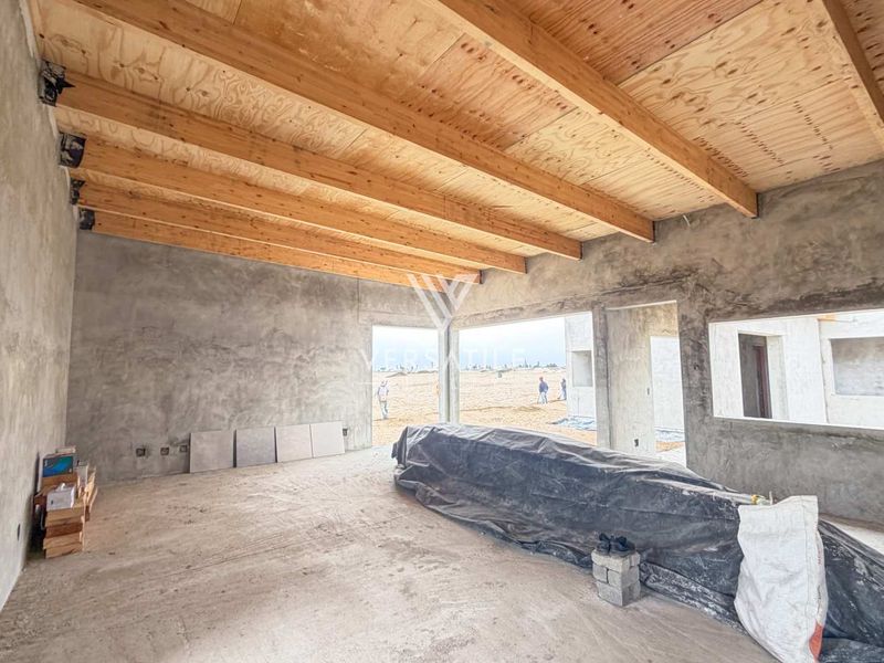 An unfinished open-plan living space featuring exposed timber beam ceilings, raw cement plaster walls, and cement screed flooring, currently under construction.