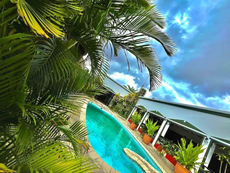 A view of the swimming pool area featuring a paved deck and adjacent covered patio space, surrounded by lush tropical landscaping.