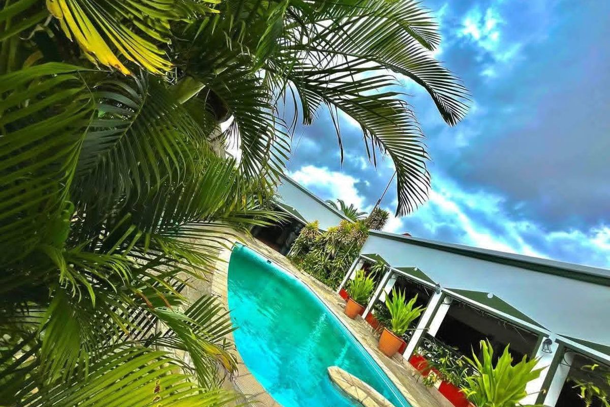 A view of the swimming pool area featuring a paved deck and adjacent covered patio space, surrounded by lush tropical landscaping.