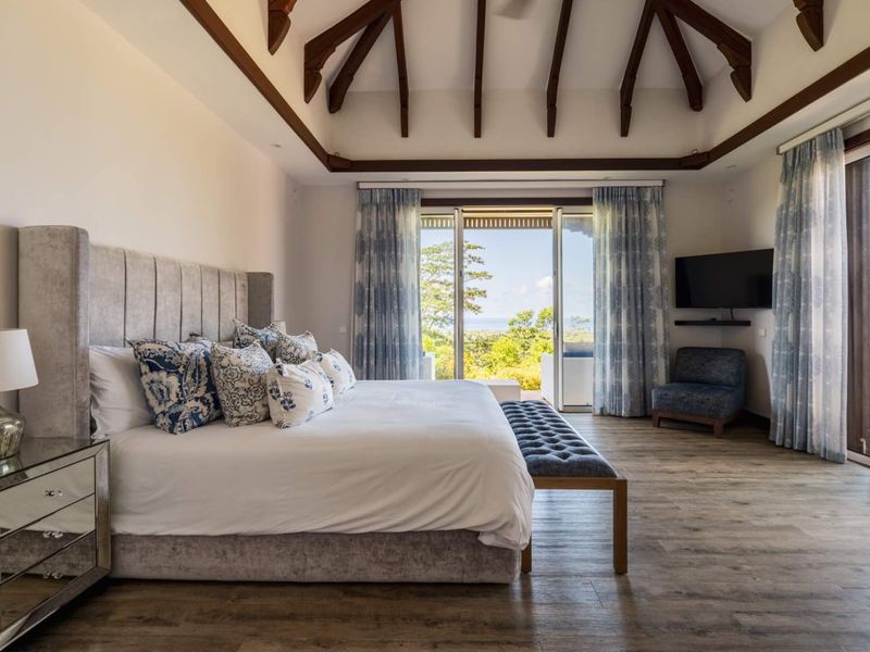 Spacious bedroom featuring high vaulted ceilings with exposed timber beams, wood-look flooring, and large sliding glass doors opening to a scenic outdoor view.