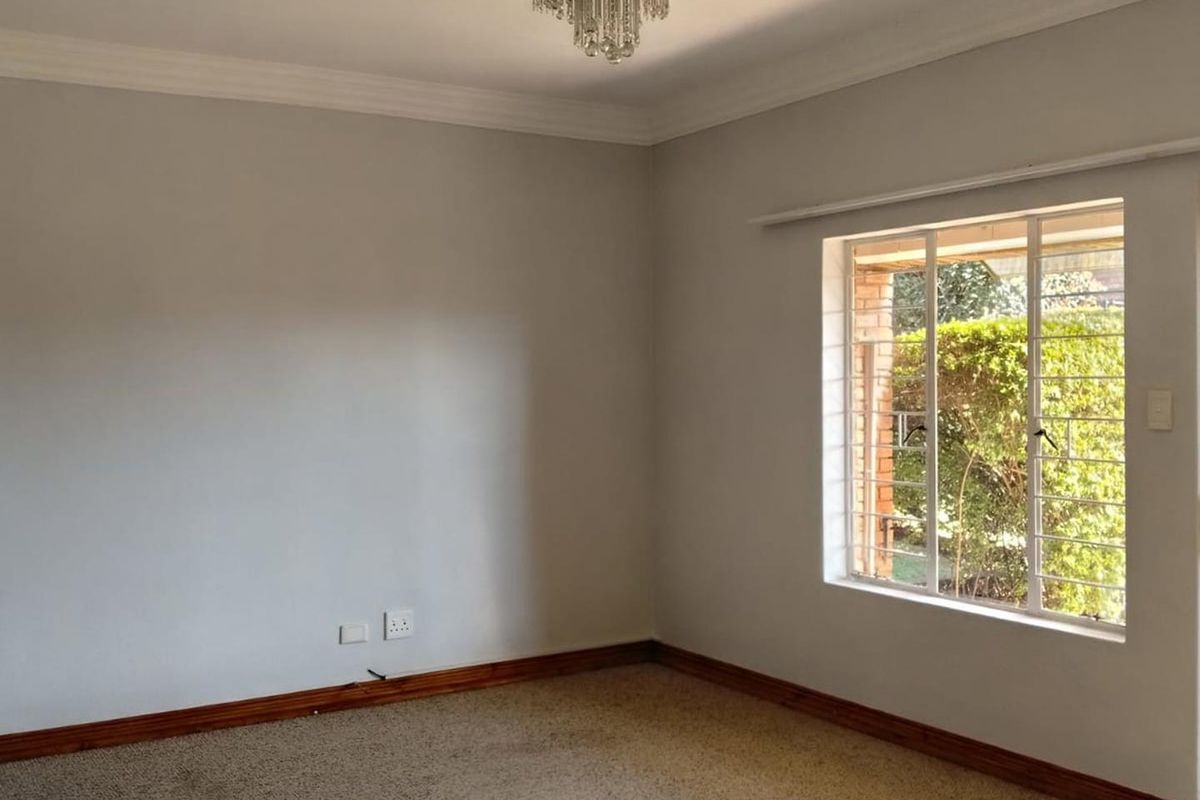 An empty bedroom featuring neutral walls, carpet flooring, timber skirting boards, and a decorative crystal chandelier, with natural light from a window with security bars.