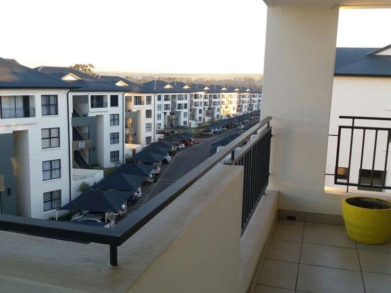 A covered balcony with tiled flooring and metal railings, offering an elevated view over the apartment complex and parking area.