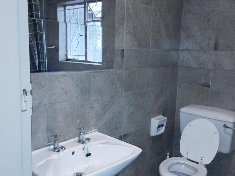 A functional bathroom featuring grey wall and floor tiles, a pedestal basin, and a standard toilet with visible plumbing.