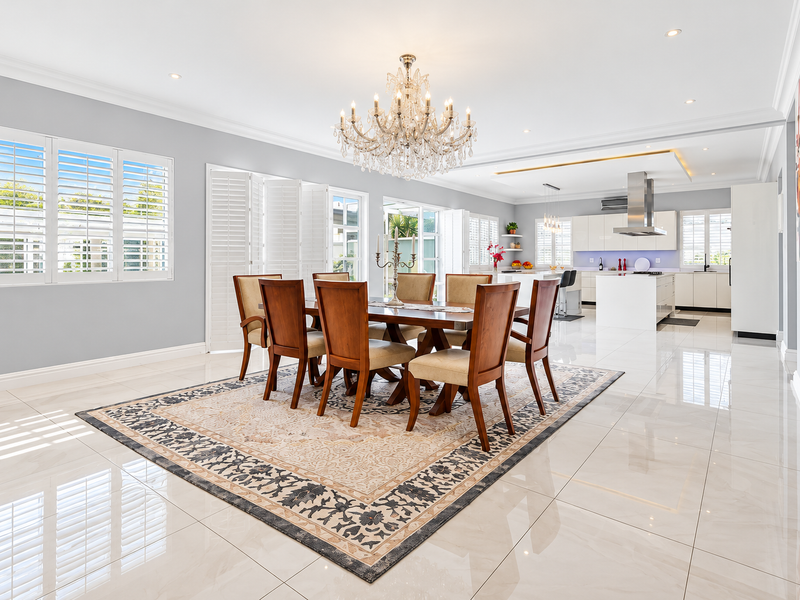 Spacious open-plan dining and kitchen area featuring polished porcelain tiles, plantation shutters, a modern kitchen island, and an elegant crystal chandelier.
