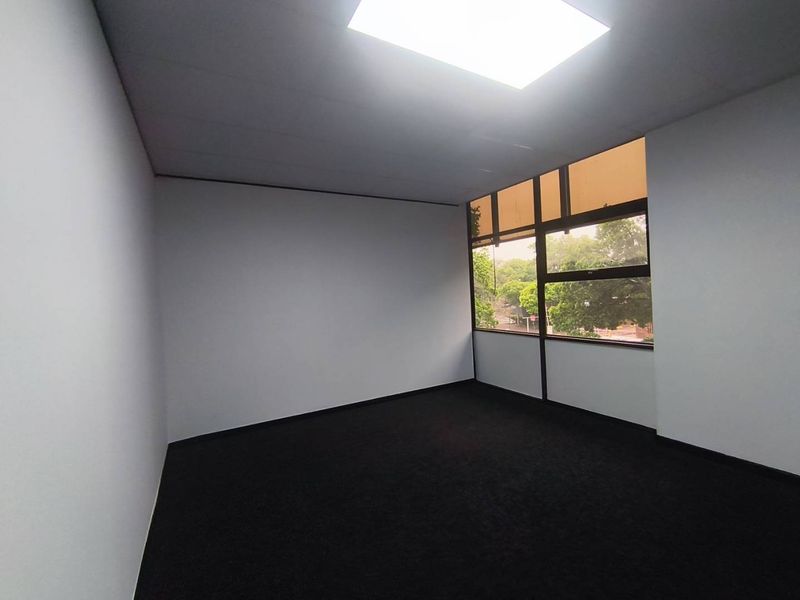 A clean, empty office space featuring dark carpeting, neutral walls, and a large window providing natural light.