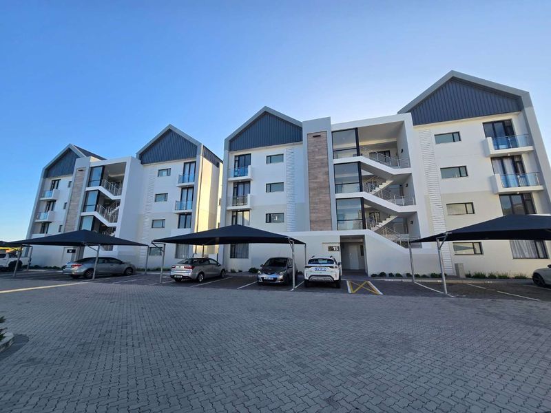 Modern apartment complex exterior featuring multi-story residential blocks with balconies, external stairwells, and dedicated covered parking bays on a paved driveway.