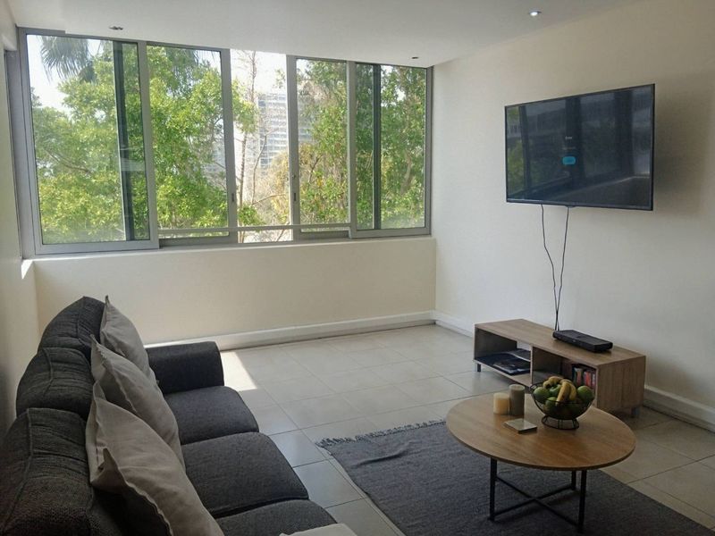 A bright living room featuring large sliding windows with a leafy outlook, neutral tiled flooring, and recessed downlighting.