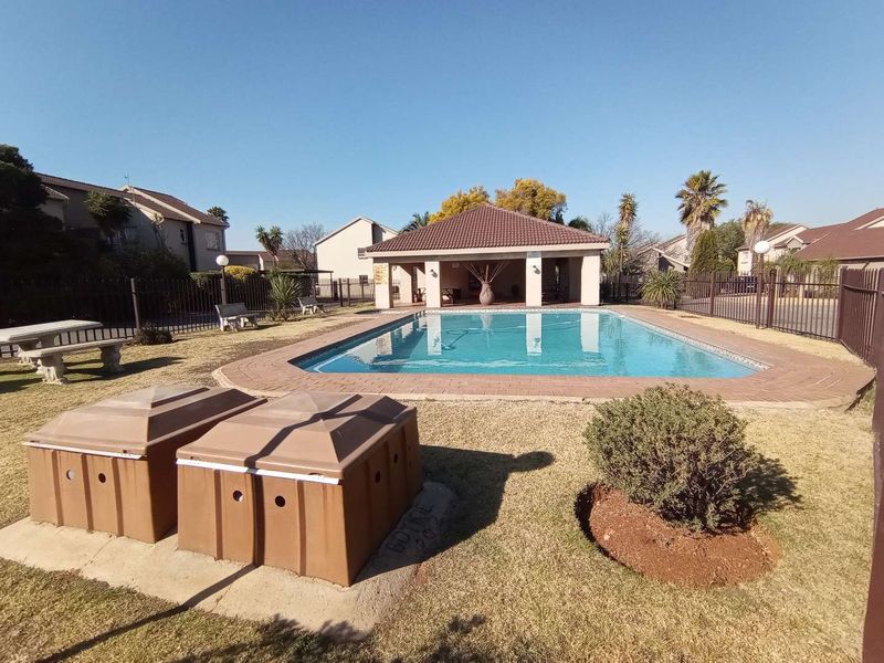 Communal swimming pool area featuring a covered patio, brick paving, and secure metal perimeter fencing within a townhouse complex.