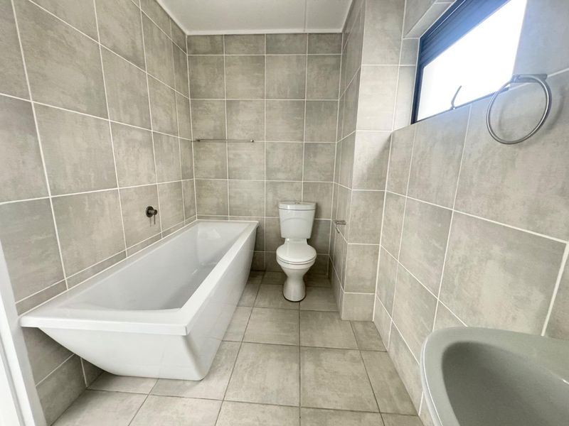 A clean, modern bathroom featuring neutral grey wall and floor tiles, a white bathtub, and a standard toilet.