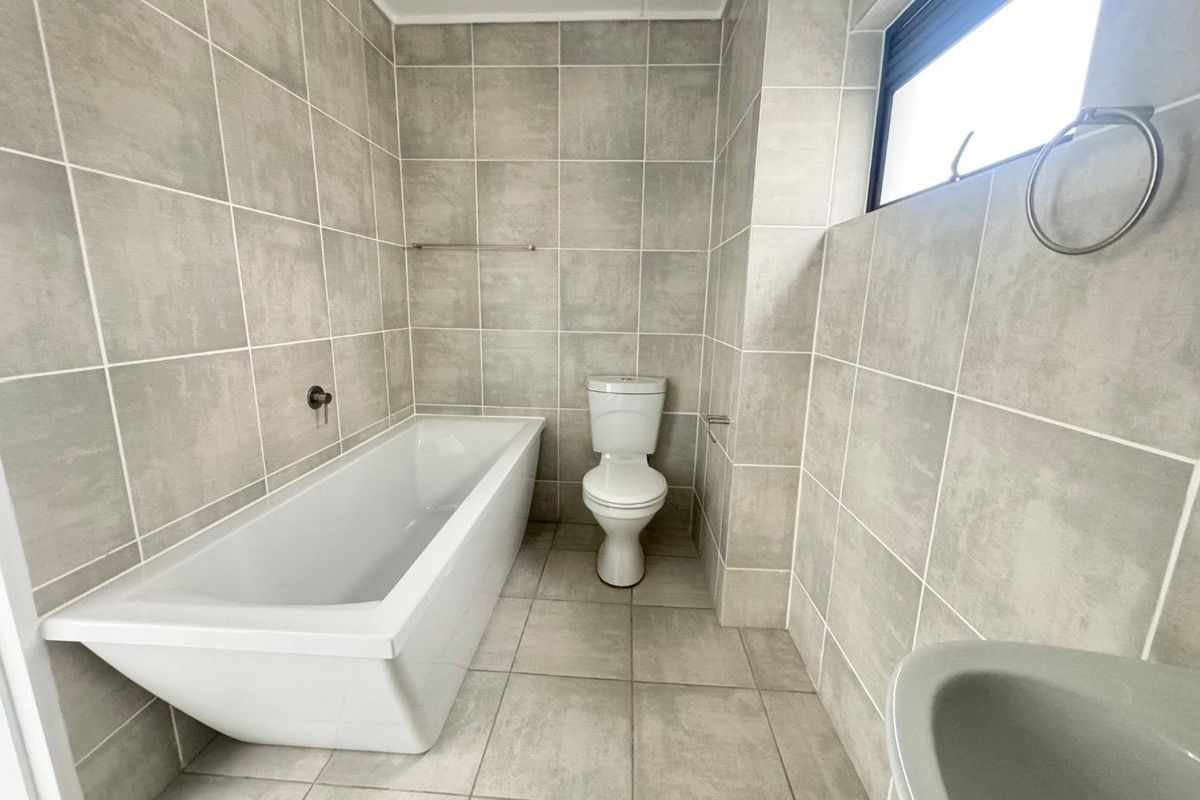 A clean, modern bathroom featuring neutral grey wall and floor tiles, a white bathtub, and a standard toilet.
