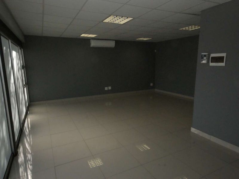 A spacious, empty office area featuring tiled flooring, a suspended ceiling with recessed lighting, and a wall-mounted air conditioning unit.