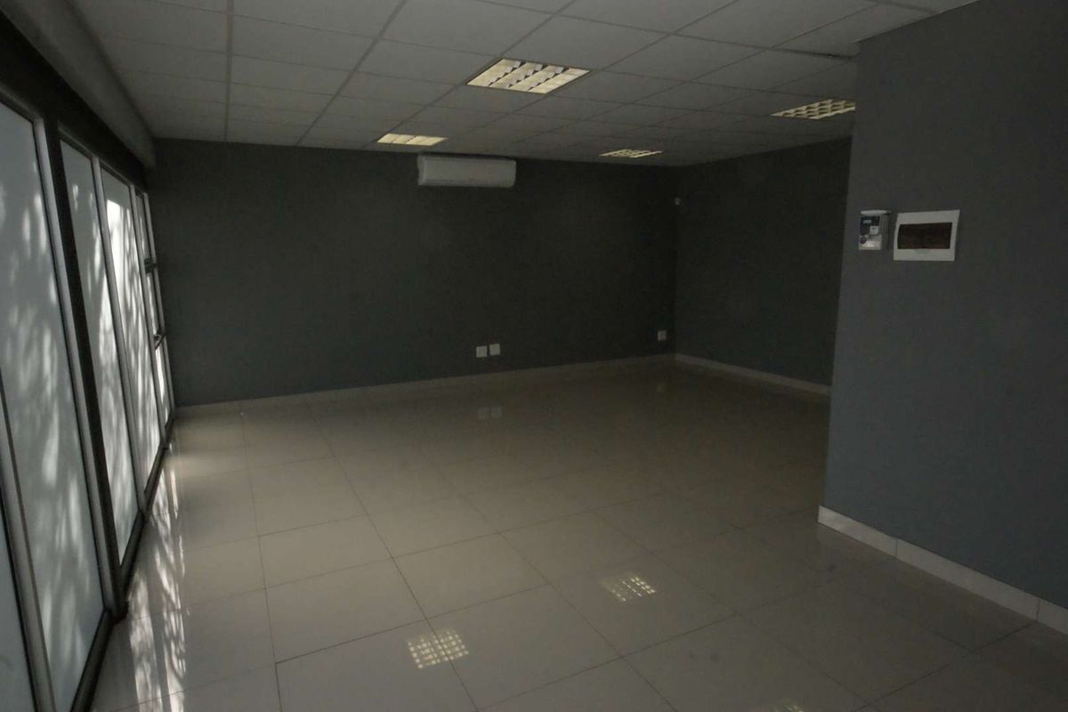 A spacious, empty office area featuring tiled flooring, a suspended ceiling with recessed lighting, and a wall-mounted air conditioning unit.