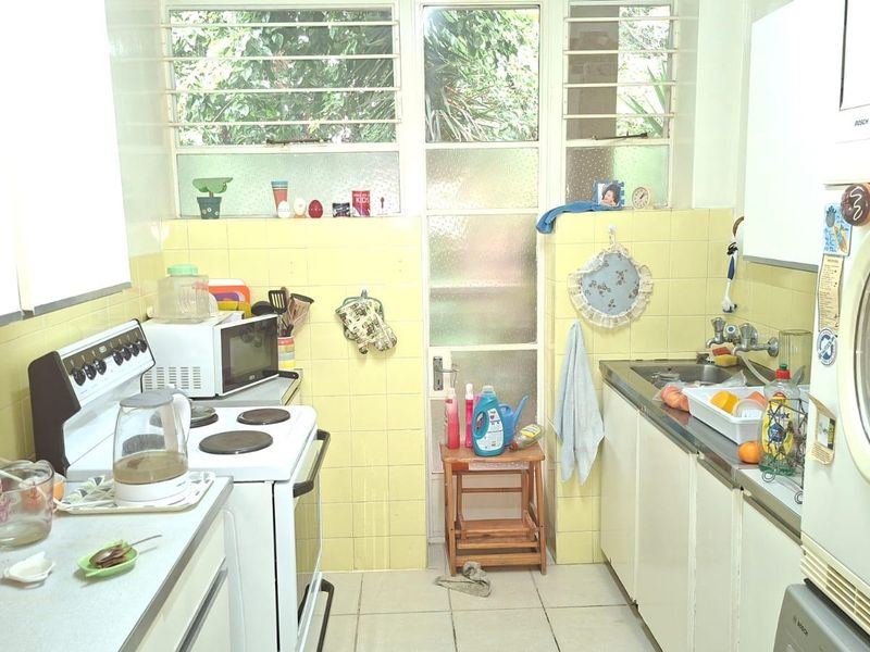 A functional kitchen featuring tiled splashbacks, built-in cabinetry, a stainless steel sink, and a freestanding stove, with louvered windows providing natural light.