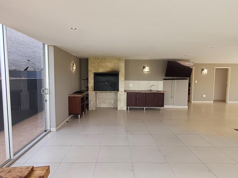 Spacious covered patio featuring a built-in braai, stone fireplace surround, and a functional kitchenette with sink, opening to an outdoor area via sliding doors.