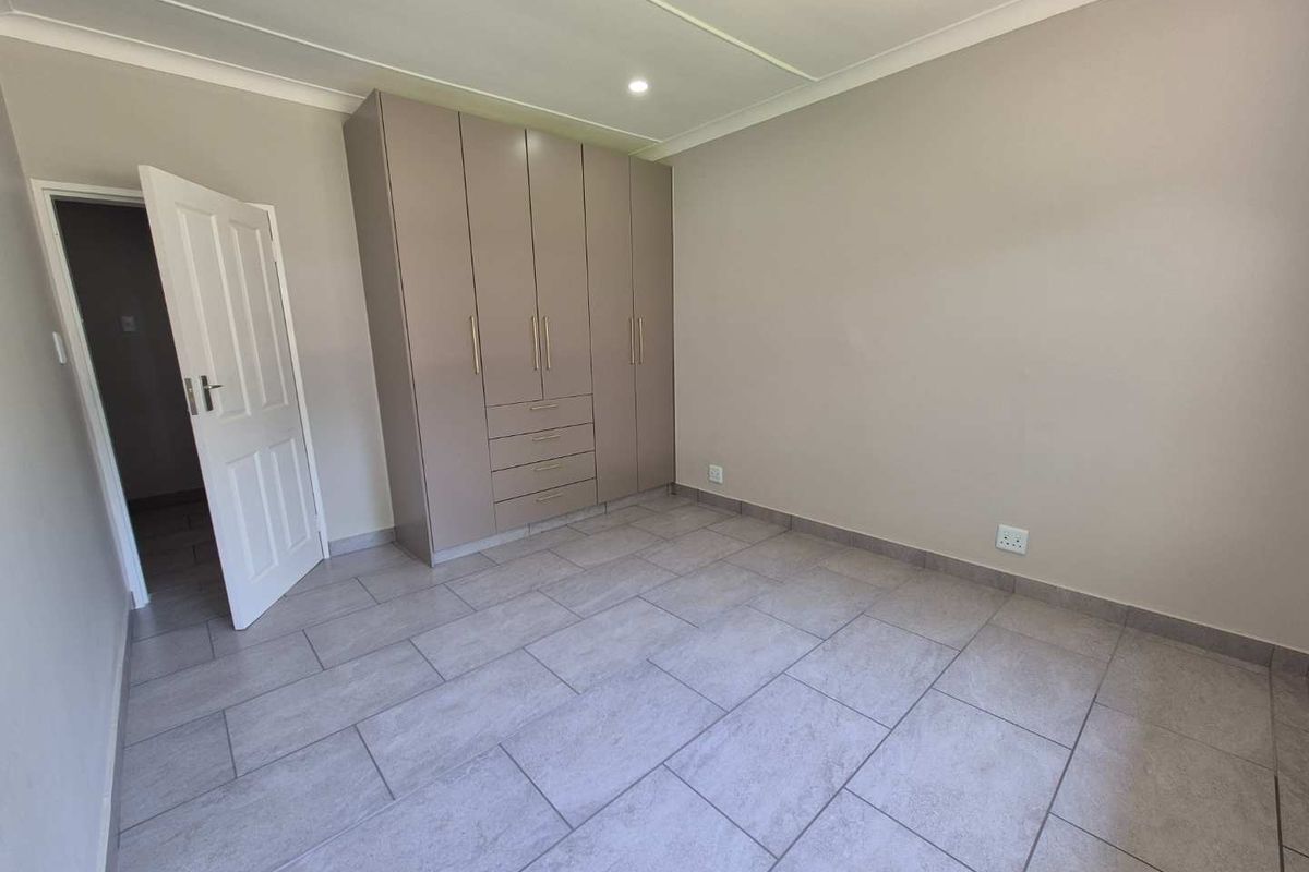 A clean, neutral bedroom featuring modern grey floor tiles, recessed ceiling lighting, and a large built-in wardrobe with integrated drawers.