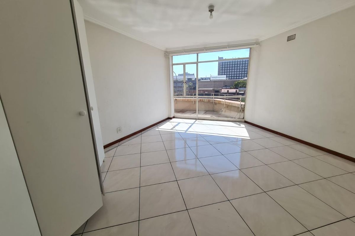 A bright, empty bedroom featuring large windows with city views, tiled flooring, and built-in cupboards.