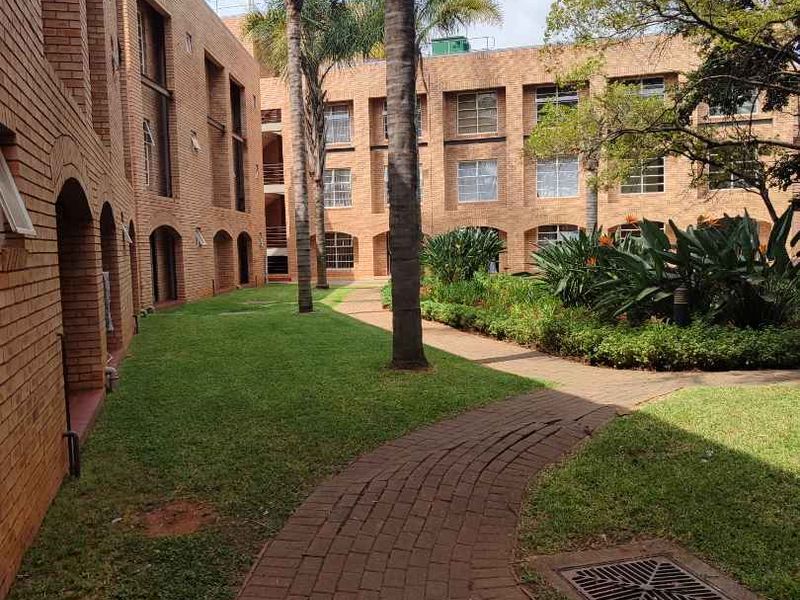 A well-maintained communal garden area featuring brick-faced apartment building exteriors, paved pathways, and established palm trees.