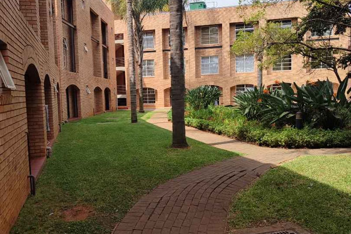 A well-maintained communal garden area featuring brick-faced apartment building exteriors, paved pathways, and established palm trees.