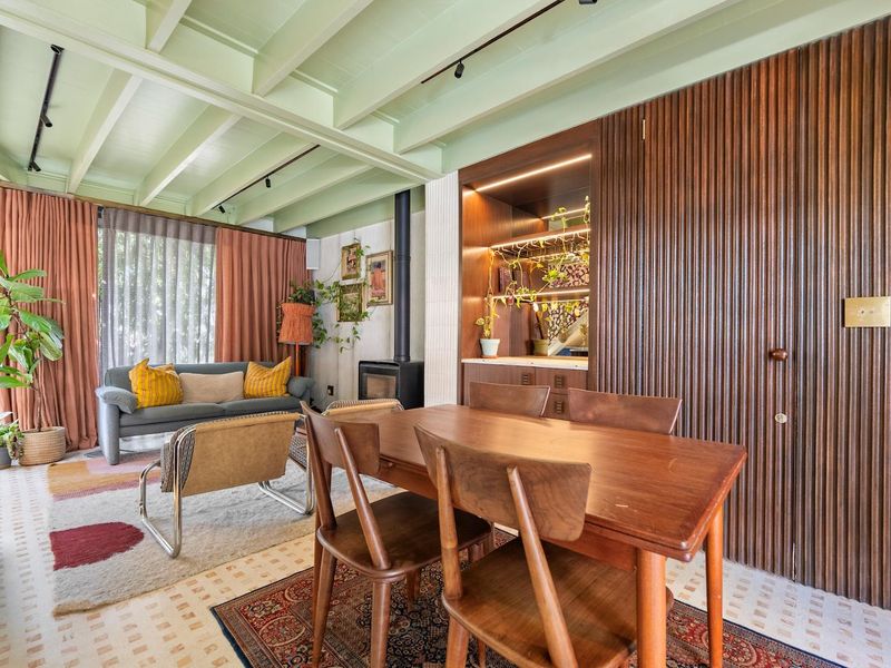 Open plan living and dining area featuring exposed beam ceilings, a wood-burning fireplace, and custom fluted wood cabinetry with integrated shelving.