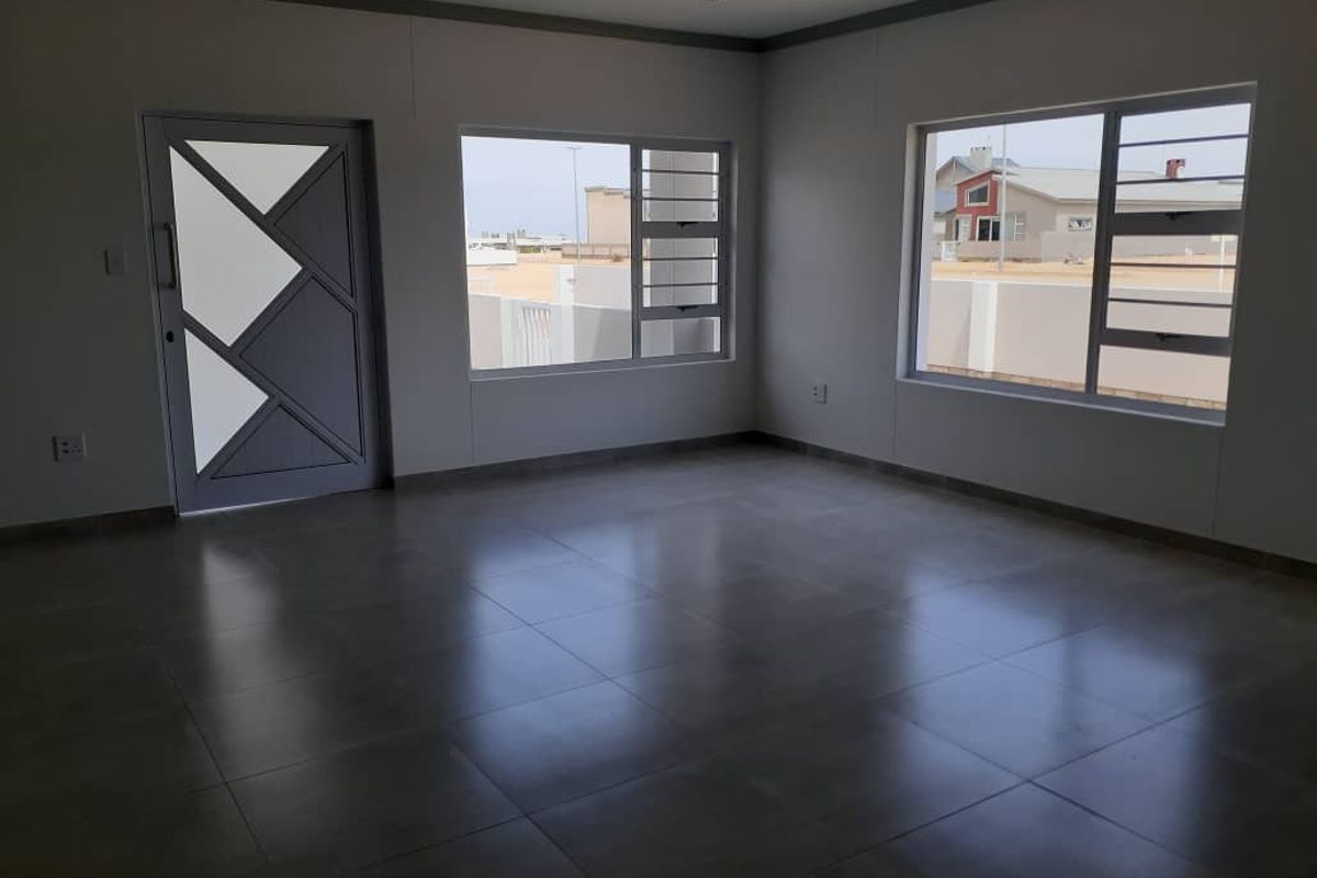 A bright, open-plan living area featuring large windows, modern grey floor tiles, and a contemporary geometric-patterned entrance door.