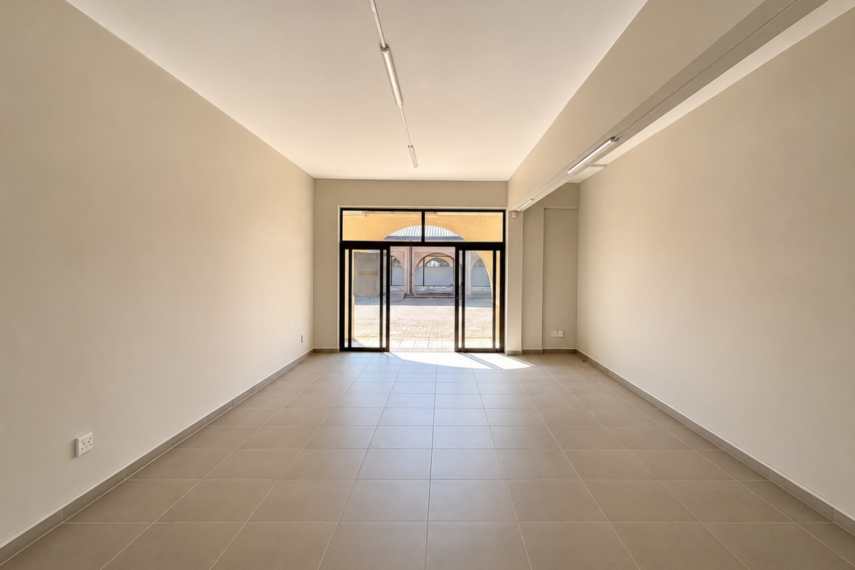 A bright, vacant open-plan retail space featuring neutral tiled flooring, clean walls, and large glass sliding doors providing natural light and exterior access.