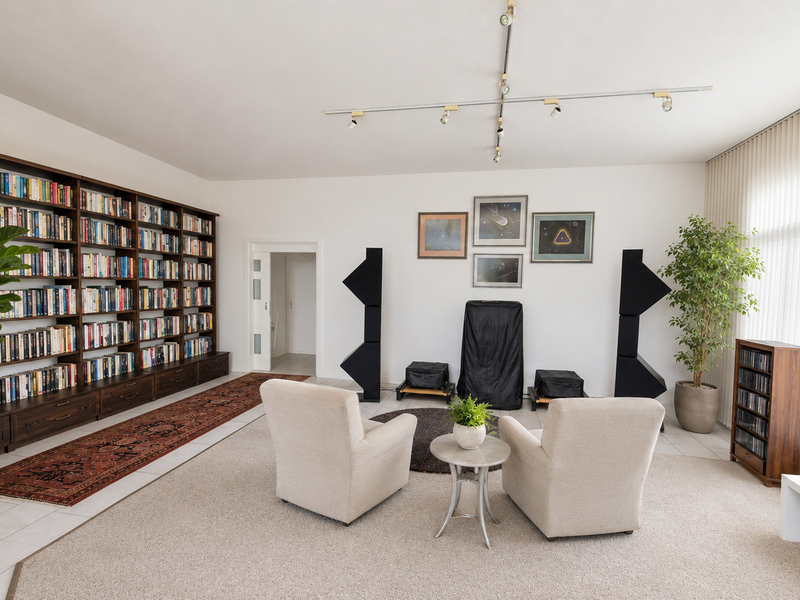 A spacious living room featuring a large built-in floor-to-ceiling bookshelf, track lighting, and expansive windows with vertical blinds.