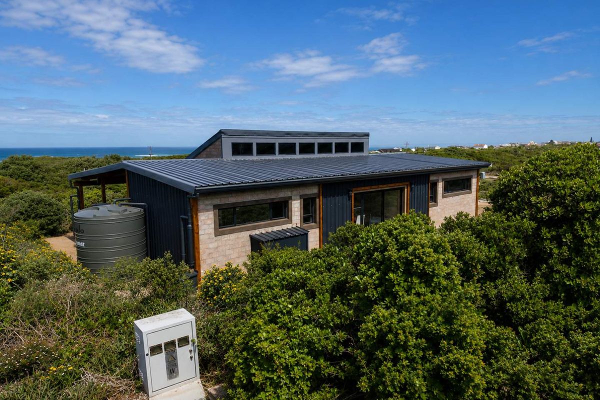 Modern coastal home featuring a metal roof, clerestory windows, and integrated water storage, set within a natural landscape with ocean views.